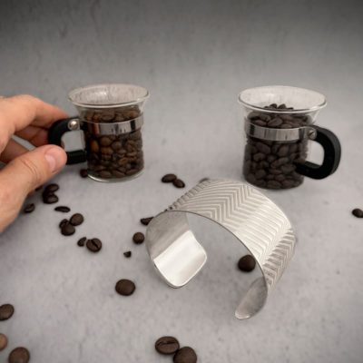 hand holding a coffee cup surrounded by coffee beans and a silver bracelet with geometric lines layered on a surface